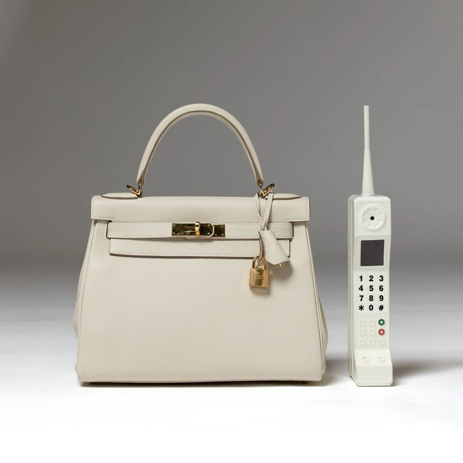 An ivory top-handle handbag with a padlock beside a white retro brick-style cordless phone on a grey background